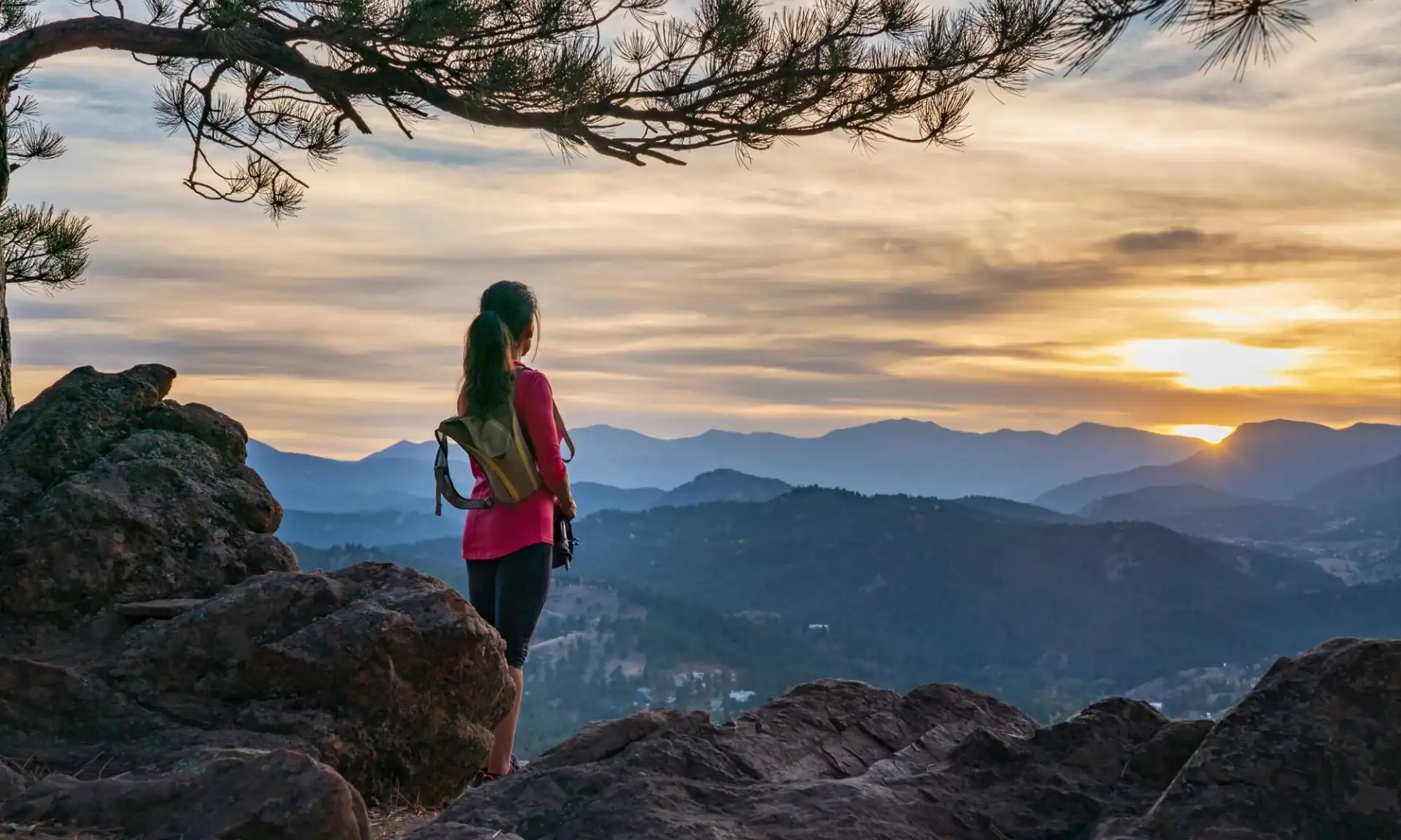 A person standing at a mountain summit at sunrise, looking out over layered Colorado ranges